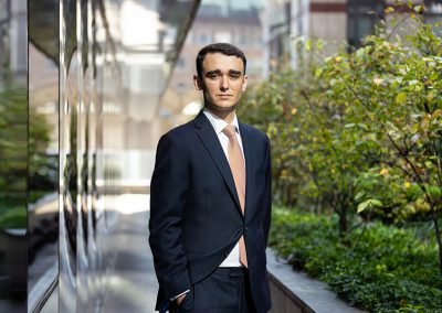 A young man in a business suite stands next to a modern office posing for a headshot