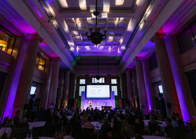 A wide angle photograph of a large pillared hall lit in purple where a conference is taking place