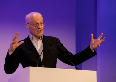 A older gentleman dramatically gestures at a podium during a corporate conference