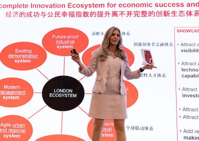 A suited woman presents at a conference, her backdrop is a digital corporate infographic